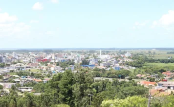 TCE suspende contrato de R$ 8,4 milhões em Sombrio Vista panorâmica em ângulo aberto da cidade de Sombrio, Santa Catarina. A imagem mostra o centro urbano com diversos edifícios, casas e ruas entrelaçadas por áreas de vegetação verde sob um céu azul com poucas nuvens. A foto ilustra o município que é alvo da fiscalização do Tribunal de Contas do Estado.