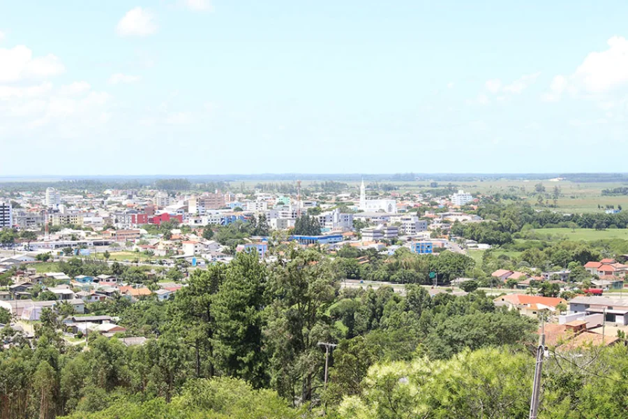Vista panorâmica em ângulo aberto da cidade de Sombrio, Santa Catarina. A imagem mostra o centro urbano com diversos edifícios, casas e ruas entrelaçadas por áreas de vegetação verde sob um céu azul com poucas nuvens. A foto ilustra o município que é alvo da fiscalização do Tribunal de Contas do Estado.