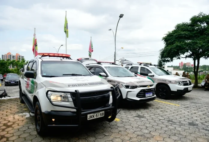Tentativa de assalto em Criciúma termina com morador ferido Três viaturas da Polícia Militar de Santa Catarina, de modelos variados e cores branca com detalhes em verde e vermelho, estão estacionadas lado a lado em um pátio. Ao fundo, é possível ver bandeiras hasteadas e parte de uma área urbana, representando a prontidão das forças de segurança que atenderam ao chamado no bairro Linha Batista.
