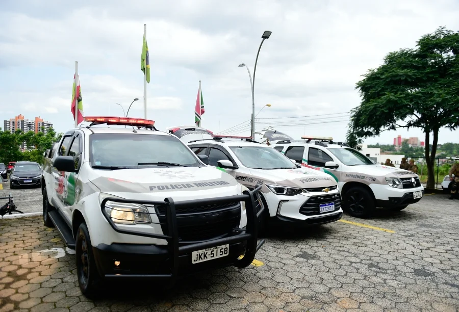 Três viaturas da Polícia Militar de Santa Catarina, de modelos variados e cores branca com detalhes em verde e vermelho, estão estacionadas lado a lado em um pátio. Ao fundo, é possível ver bandeiras hasteadas e parte de uma área urbana, representando a prontidão das forças de segurança que atenderam ao chamado no bairro Linha Batista.