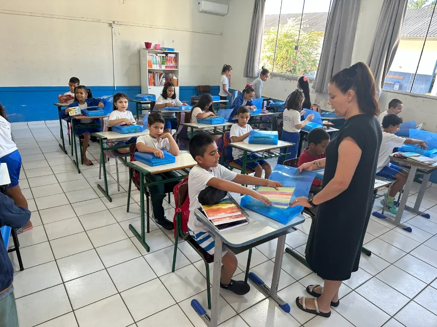 Alunos da EMEF Frei Modesto recebem materiais escolares no primeiro dia de aula. A imagem mostra as crianças entusiasmadas manuseando os kits sobre as mesas enquanto a professora orienta a atividade.