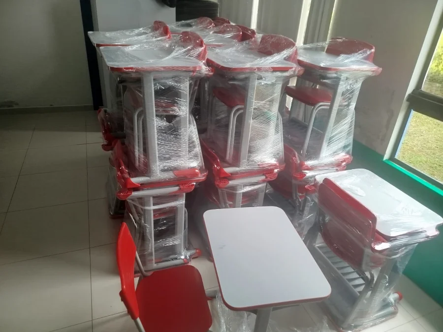 A imagem mostra várias pilhas de conjuntos escolares novos, compostos por carteiras e cadeiras nas cores vermelho e branco. Os móveis estão embalados em plástico transparente de proteção e organizados dentro de uma sala iluminada, simbolizando a renovação do mobiliário para a rede municipal de ensino de Timbé do Sul.