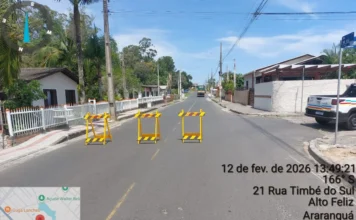 Trânsito sofre alteração no Alto Feliz em Araranguá A imagem exibe uma vista em perspectiva da Rua Timbé do Sul, no bairro Alto Feliz, em Araranguá. O asfalto está seco e a via encontra-se bloqueada por três cavaletes de sinalização amarelos posicionados lado a lado, impedindo a passagem de veículos. Ao fundo, observa-se o ambiente residencial com casas, muros brancos e vegetação. No canto inferior esquerdo, há um detalhe do mapa da região e, no lado direito, informações técnicas sobre a localização, data (12 de fevereiro de 2026) e coordenadas geográficas, reforçando o registro oficial da interdição para as obras de drenagem.