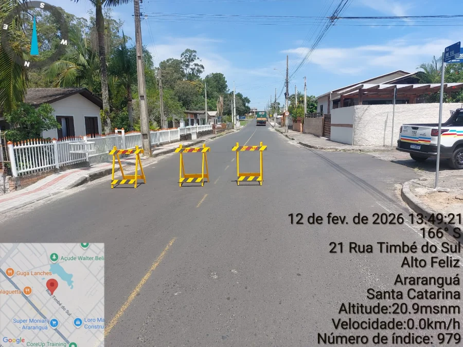 A imagem exibe uma vista em perspectiva da Rua Timbé do Sul, no bairro Alto Feliz, em Araranguá. O asfalto está seco e a via encontra-se bloqueada por três cavaletes de sinalização amarelos posicionados lado a lado, impedindo a passagem de veículos. Ao fundo, observa-se o ambiente residencial com casas, muros brancos e vegetação. No canto inferior esquerdo, há um detalhe do mapa da região e, no lado direito, informações técnicas sobre a localização, data (12 de fevereiro de 2026) e coordenadas geográficas, reforçando o registro oficial da interdição para as obras de drenagem.
