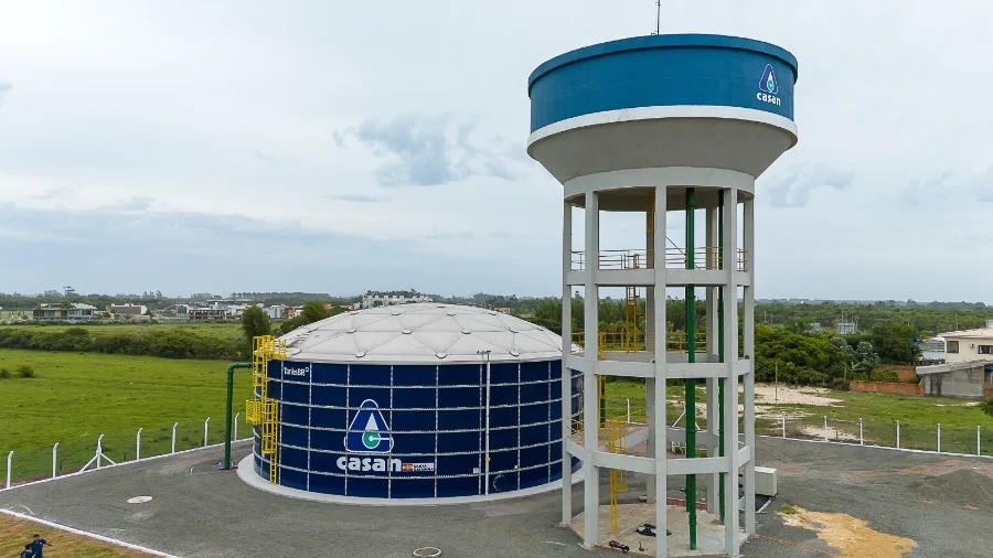 Última semana para quitar débitos com a CASAN com desconto Fotografia aérea de uma unidade operacional da CASAN, apresentando em primeiro plano um grande reservatório cilíndrico de cor azul escura com o logotipo da companhia e, ao lado, uma torre de água elevada em concreto. A estrutura está situada em uma área gramada sob um céu nublado, representando a infraestrutura de saneamento da Companhia em Santa Catarina.
