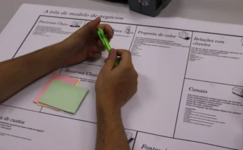 Unesc abre inscrições para consultoria gratuita a empresas Fotografia em close foca nas mãos de um acadêmico segurando uma caneta verde sobre um cartaz de "Tela de modelo de negócios" (Business Model Canvas). Sobre o papel, há blocos de notas adesivas nas cores rosa e verde. A imagem ilustra o processo de análise estratégica e diagnóstico empresarial realizado durante o programa Plano 60 Dias.