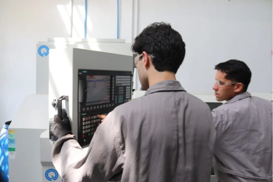 Dois estudantes dos cursos de Engenharia e Tecnologia da Unesc, usando óculos de proteção e macacões cinza, operam uma máquina industrial com painel de controle digital em um laboratório da universidade, ilustrando a formação prática oferecida.
