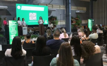 Unesc abre vagas para curso inédito de Terapia Ocupacional Fotografia de um evento acadêmico realizado em um auditório da Unesc. No palco, uma mulher vestindo calça marrom e camiseta branca fala ao microfone durante a Aula Magna do novo curso de Terapia Ocupacional. À esquerda, outras três mulheres acompanham a apresentação ao lado de um banner informativo verde. Ao fundo, um grande telão digital exibe um QR Code sob o título do curso. No primeiro plano, o público é visto de costas, sentado em cadeiras pretas, acompanhando a palestra.