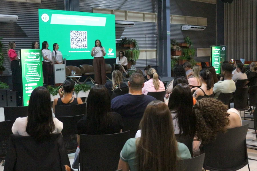 Fotografia de um evento acadêmico realizado em um auditório da Unesc. No palco, uma mulher vestindo calça marrom e camiseta branca fala ao microfone durante a Aula Magna do novo curso de Terapia Ocupacional. À esquerda, outras três mulheres acompanham a apresentação ao lado de um banner informativo verde. Ao fundo, um grande telão digital exibe um QR Code sob o título do curso. No primeiro plano, o público é visto de costas, sentado em cadeiras pretas, acompanhando a palestra.
