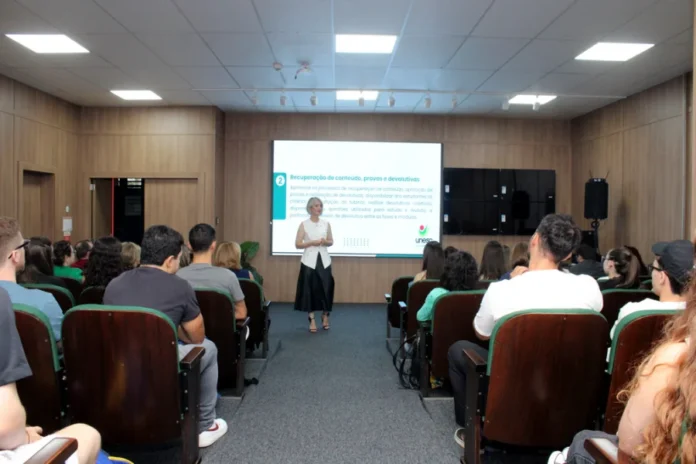 Unesc alinha nova matriz pedagógica com alunos de Medicina Fotografia em plano médio de um auditório na Unesc. No centro, a reitora em exercício, Gisele Coelho Lopes, está de pé falando aos estudantes. Ela possui cabelos curtos e loiros, veste um colete branco e saia longa preta. Ao fundo, um telão exibe um slide com o logotipo da Unesc e o título "Recuperação de conteúdo, provas e devolutivas", detalhando tópicos da nova Matriz 6. Em primeiro plano, as costas de diversos acadêmicos de Medicina sentados em poltronas de madeira, acompanhando a apresentação.