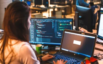 Unesc amplia oferta de curso de Inteligência Artificial Mulher em um ambiente tecnológico moderno, vista de costas e lateralmente, trabalhando concentrada em um notebook e em um monitor grande que exibe várias linhas de código colorido. A imagem ilustra a aprendizagem prática de programação e o desenvolvimento de algoritmos, focos do novo curso Superior de Tecnologia em Inteligência Artificial da Unesc.