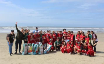 Unesc aprova projeto ambiental em rede internacional Integrantes do projeto de extensão da Unesc focado em resíduos marinhos sorriem para a câmera em uma faixa de areia. A imagem destaca a ação prática de limpeza costeira, com sacos de lixo em primeiro plano e o mar ao fundo.