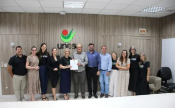 Unesc e Sicredi firmam parceria pelo turismo regional Representantes da Unesc e do Sicredi Sul SC posam perfilados para foto na reitoria da Universidade. No centro do grupo, a reitora em exercício, Gisele Silveira Coelho Lopes, e o presidente do Sicredi Sul SC, Aloísio Westrup, seguram juntos o contrato assinado da parceria para o programa Integratur. O grupo está em uma sala com painéis de madeira ao fundo e o logotipo verde e vermelho da Unesc centralizado na parede.