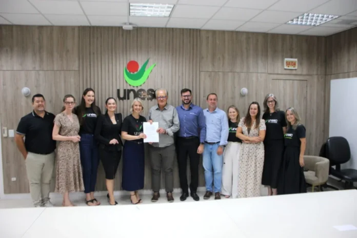 Unesc e Sicredi firmam parceria pelo turismo regional Representantes da Unesc e do Sicredi Sul SC posam perfilados para foto na reitoria da Universidade. No centro do grupo, a reitora em exercício, Gisele Silveira Coelho Lopes, e o presidente do Sicredi Sul SC, Aloísio Westrup, seguram juntos o contrato assinado da parceria para o programa Integratur. O grupo está em uma sala com painéis de madeira ao fundo e o logotipo verde e vermelho da Unesc centralizado na parede.