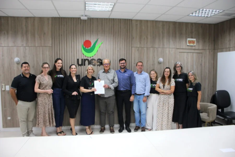 Representantes da Unesc e do Sicredi Sul SC posam perfilados para foto na reitoria da Universidade. No centro do grupo, a reitora em exercício, Gisele Silveira Coelho Lopes, e o presidente do Sicredi Sul SC, Aloísio Westrup, seguram juntos o contrato assinado da parceria para o programa Integratur. O grupo está em uma sala com painéis de madeira ao fundo e o logotipo verde e vermelho da Unesc centralizado na parede.