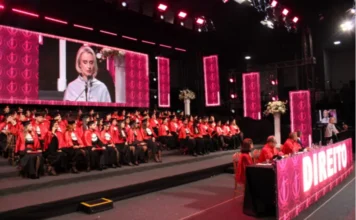 Unesc forma nova turma de Direito em solenidade festiva Fotografia de ângulo aberto da cerimônia de colação de grau do curso de Direito da Unesc, realizada no salão AM Master Hall. À esquerda, dezenas de formandos vestem becas pretas com faixas vermelhas e capelos, sentados em degraus. Ao fundo, um grande telão exibe em destaque o rosto da reitora em exercício, Gisele Silveira Coelho Lopes, durante seu pronunciamento. À direita, autoridades acadêmicas compõem a mesa de honra, decorada com a palavra "DIREITO" em letras brancas iluminadas sobre um painel rosa.