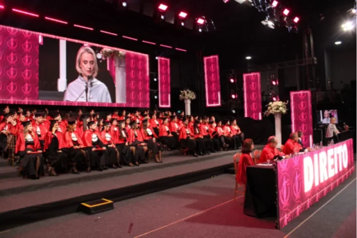 Unesc forma nova turma de Direito em solenidade festiva Fotografia de ângulo aberto da cerimônia de colação de grau do curso de Direito da Unesc, realizada no salão AM Master Hall. À esquerda, dezenas de formandos vestem becas pretas com faixas vermelhas e capelos, sentados em degraus. Ao fundo, um grande telão exibe em destaque o rosto da reitora em exercício, Gisele Silveira Coelho Lopes, durante seu pronunciamento. À direita, autoridades acadêmicas compõem a mesa de honra, decorada com a palavra "DIREITO" em letras brancas iluminadas sobre um painel rosa.