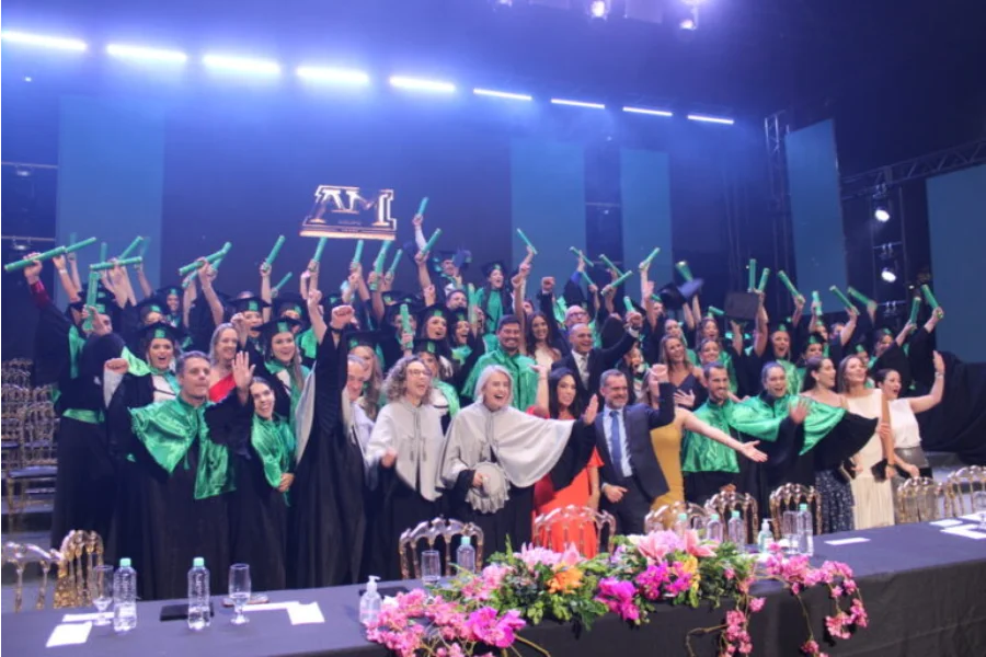 Formandos dos cursos de Farmácia, Fisioterapia e Tecnologia em Estética e Cosmética da Unesc celebram no palco do AM Master Hall. O grupo veste becas pretas com faixas e capelos verdes, erguendo com entusiasmo seus diplomas. Ao fundo, luzes de palco azuis iluminam o ambiente, e à frente, uma mesa decorada com flores rosas e brancas compõe o cenário da solenidade.