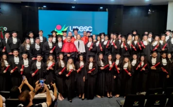 Unesc forma novos profissionais em cerimônia de gestão Grande grupo de formandos da universidade Unesc reunidos em um palco de auditório, vestindo becas pretas e segurando canudos vermelhos de diploma. Ao centro, autoridades acadêmicas usam vestes diferenciadas e, ao fundo, um telão exibe o logotipo da instituição. Na plateia, pessoas registram o momento com celulares.
