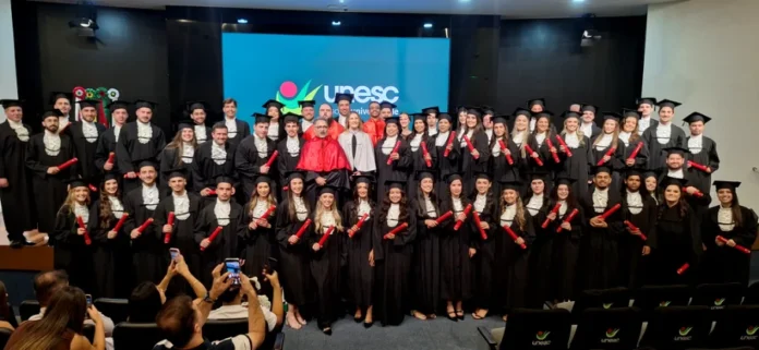 Unesc forma novos profissionais em cerimônia de gestão Grande grupo de formandos da universidade Unesc reunidos em um palco de auditório, vestindo becas pretas e segurando canudos vermelhos de diploma. Ao centro, autoridades acadêmicas usam vestes diferenciadas e, ao fundo, um telão exibe o logotipo da instituição. Na plateia, pessoas registram o momento com celulares.