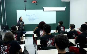 Unesc inicia ano letivo 2026 com foco no magistério Foto de uma sala de aula da Unesc, com uma professora em pé, escrevendo no quadro branco sobre 'História da Arte'. Em frente a ela, vários alunos estão sentados em carteiras, assistindo à aula. Todos os alunos vestem camisetas pretas com uma estampa vermelha e branca nas costas que diz 'TERCEIRÃO 2K26'. A imagem ilustra o ambiente acadêmico e a formação de estudantes, temas centrais do Programa Nossa Bolsa.