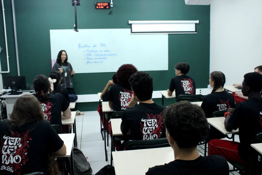 Foto de uma sala de aula da Unesc, com uma professora em pé, escrevendo no quadro branco sobre 'História da Arte'. Em frente a ela, vários alunos estão sentados em carteiras, assistindo à aula. Todos os alunos vestem camisetas pretas com uma estampa vermelha e branca nas costas que diz 'TERCEIRÃO 2K26'. A imagem ilustra o ambiente acadêmico e a formação de estudantes, temas centrais do Programa Nossa Bolsa.