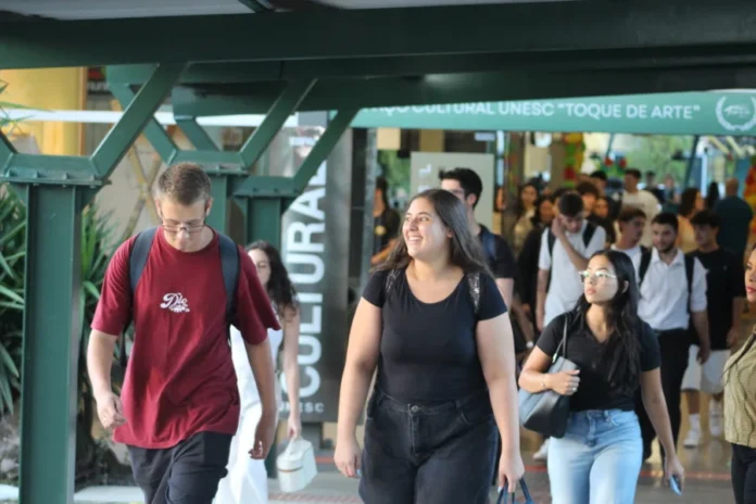 Unesc inicia letivo 2026 com foco em diversidade e inclusão Estudantes de diversos perfis caminham pelo Espaço Cultural "Toque de Arte" no campus da Unesc, em Criciúma. No centro da imagem, uma jovem sorri enquanto transita pelo local, representando a movimentação e o clima de acolhimento no primeiro dia de aulas do semestre letivo de 2026.