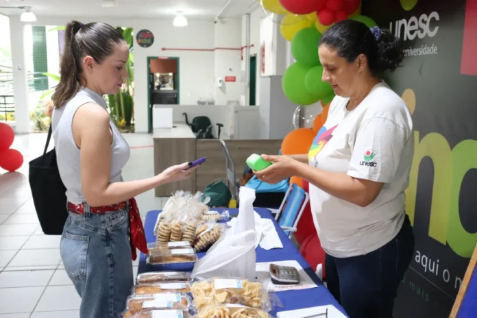 Unesc retoma Feira da Economia Solidária no campus Uma produtora local e uma consumidora interagem em uma das bancas da Feira da Economia Solidária no campus da Unesc. A cliente, à esquerda, utiliza o celular para realizar um pagamento, enquanto a feirante, vestindo uma camiseta branca com o logotipo da universidade, organiza produtos embalados sobre uma mesa com toalha azul. Na banca, são vistos diversos pacotes de pães e biscoitos artesanais. Ao fundo, balões coloridos decoram o ambiente interno do bloco.