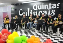 Unesc retoma Quintas Culturais com foco em inclusão Grupo musical de samba e pagode se apresenta no palco do projeto Quintas Culturais da Unesc. Os músicos tocam instrumentos como pandeiro, violão, cavaquinho e trompete. À frente do palco, há balões coloridos e, entre os integrantes, uma criança pequena participa da apresentação, reforçando o caráter inclusivo do evento.