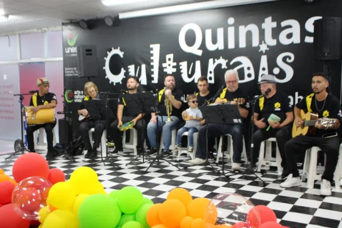 Unesc retoma Quintas Culturais com foco em inclusão Grupo musical de samba e pagode se apresenta no palco do projeto Quintas Culturais da Unesc. Os músicos tocam instrumentos como pandeiro, violão, cavaquinho e trompete. À frente do palco, há balões coloridos e, entre os integrantes, uma criança pequena participa da apresentação, reforçando o caráter inclusivo do evento.