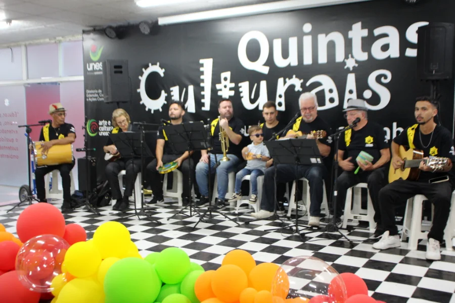 Grupo musical de samba e pagode se apresenta no palco do projeto Quintas Culturais da Unesc. Os músicos tocam instrumentos como pandeiro, violão, cavaquinho e trompete. À frente do palco, há balões coloridos e, entre os integrantes, uma criança pequena participa da apresentação, reforçando o caráter inclusivo do evento.