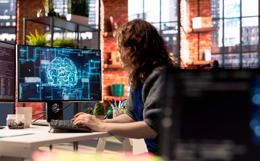 Mulher em ambiente de escritório moderno, trabalhando em um computador com uma representação gráfica de um cérebro humano feito de circuitos digitais e a sigla 