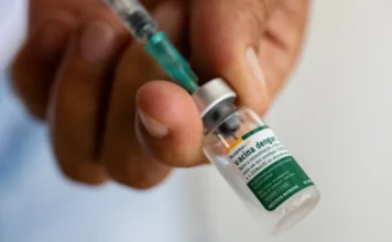 Vacina do Butantan contra dengue chega aos brasileiros Close-up de uma mão segurando um pequeno frasco de vidro da vacina contra dengue Butantan-DV, com uma seringa inserida na rolha. O frasco contém o imunizante tetravalente desenvolvido pelo Instituto Butantan para proteger contra os quatro sorotipos do vírus da dengue (DENV-1, DENV-2, DENV-3 e DENV-4). O rótulo "Butantan-DV" e "Vacina dengue" está visível no frasco.