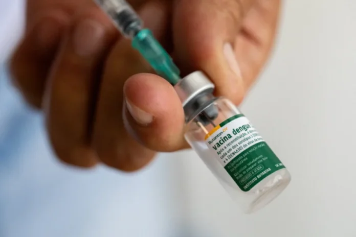 Vacina do Butantan contra dengue chega aos brasileiros Close-up de uma mão segurando um pequeno frasco de vidro da vacina contra dengue Butantan-DV, com uma seringa inserida na rolha. O frasco contém o imunizante tetravalente desenvolvido pelo Instituto Butantan para proteger contra os quatro sorotipos do vírus da dengue (DENV-1, DENV-2, DENV-3 e DENV-4). O rótulo "Butantan-DV" e "Vacina dengue" está visível no frasco.