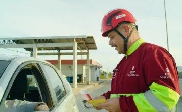 ViaCosteira reforça segurança na BR-101 Sul para o Carnaval Colaborador da ViaCosteira, identificado como Anderson e usando uniforme e capacete de segurança, entrega um folheto educativo a um motorista dentro de um carro prata. A ação de conscientização sobre segurança viária ocorre em um ponto de apoio ao longo da rodovia BR-101 Sul, em um dia ensolarado.