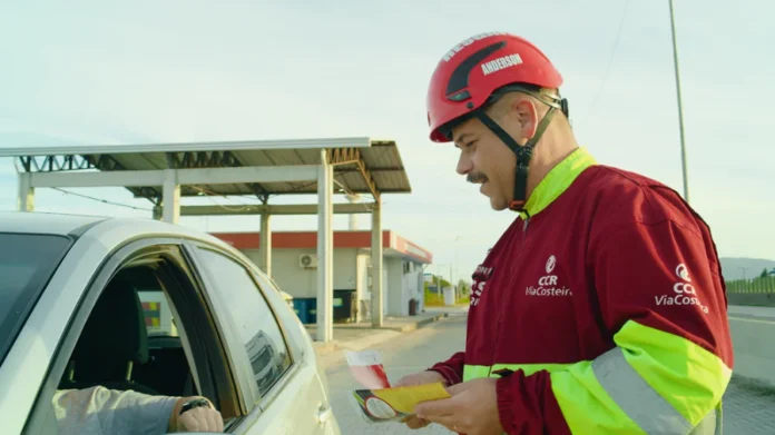 ViaCosteira reforça segurança na BR-101 Sul para o Carnaval Colaborador da ViaCosteira, identificado como Anderson e usando uniforme e capacete de segurança, entrega um folheto educativo a um motorista dentro de um carro prata. A ação de conscientização sobre segurança viária ocorre em um ponto de apoio ao longo da rodovia BR-101 Sul, em um dia ensolarado.