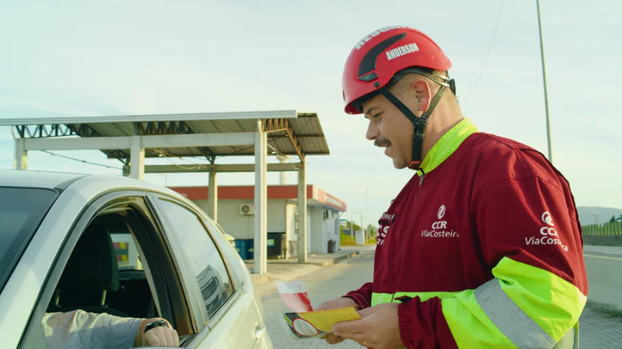 ViaCosteira reforça segurança na BR-101 Sul para o Carnaval Colaborador da ViaCosteira, identificado como Anderson e usando uniforme e capacete de segurança, entrega um folheto educativo a um motorista dentro de um carro prata. A ação de conscientização sobre segurança viária ocorre em um ponto de apoio ao longo da rodovia BR-101 Sul, em um dia ensolarado.