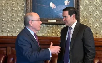 Vieira e Rubio alinham visita de Lula aos EUA em março Fotografia em plano médio do Ministro das Relações Exteriores, Mauro Vieira, e do Secretário de Estado dos EUA, Marco Rubio. Vieira, à esquerda, gesticula enquanto fala, e Rubio, à direita, escuta atentamente. Ambos vestem ternos escuros e gravatas em tons de vinho e roxo. Eles estão em uma sala de reuniões formal com papel de parede ornamentado e uma pintura clássica ao fundo.