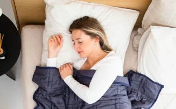 Vigitel: 20% dos adultos dormem menos de 6 horas por noite Fotografia aérea de uma mulher jovem de pele clara, deitada de lado em uma cama com lençóis e travesseiros brancos, dormindo tranquilamente sob um edredom azul escuro. A imagem ilustra o estado de repouso ideal e a importância da higiene do sono mencionada na pesquisa do Ministério da Saúde.