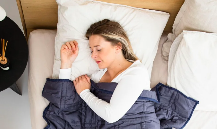 Vigitel 20% dos adultos dormem menos de 6 horas por noite Fotografia aérea de uma mulher jovem de pele clara, deitada de lado em uma cama com lençóis e travesseiros brancos, dormindo tranquilamente sob um edredom azul escuro. A imagem ilustra o estado de repouso ideal e a importância da higiene do sono mencionada na pesquisa do Ministério da Saúde.