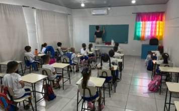 Volta às aulas em Meleiro reúne 1.100 alunos nesta segunda Fotografia do interior de uma sala de aula iluminada durante o retorno às aulas em Meleiro. Uma professora, de costas, escreve no quadro negro enquanto cerca de 15 alunos crianças, vestindo uniformes brancos e azuis, observam sentados em suas carteiras organizadas em fileiras. A sala possui paredes brancas, ar-condicionado instalado e janelas com cortinas claras, além de uma cortina colorida em tons de arco-íris ao fundo.