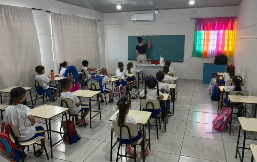 Volta às aulas em Meleiro reúne 1.100 alunos nesta segunda Fotografia do interior de uma sala de aula iluminada durante o retorno às aulas em Meleiro. Uma professora, de costas, escreve no quadro negro enquanto cerca de 15 alunos crianças, vestindo uniformes brancos e azuis, observam sentados em suas carteiras organizadas em fileiras. A sala possui paredes brancas, ar-condicionado instalado e janelas com cortinas claras, além de uma cortina colorida em tons de arco-íris ao fundo.