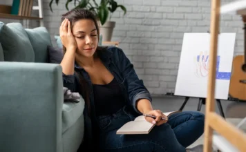 A importância de priorizar a saúde mental no dia a dia Uma jovem sentada no chão, com a mão na cabeça e os olhos fechados, demonstrando cansaço e introspecção. Ela segura um caderno e caneta, com elementos criativos como um cavalete de arte e um violão ao fundo, ilustrando a necessidade de pausa e autocuidado em meio a uma rotina agitada, conforme discutido na matéria sobre saúde mental.