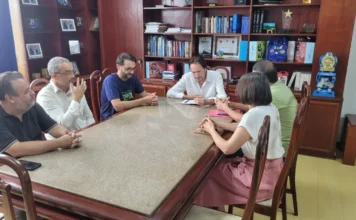 ACIVA e UFSC buscam retomar obra do Centro de Saúde Fotografia colorida em plano médio de uma reunião institucional realizada em um gabinete oficial. Seis pessoas, cinco homens e uma mulher, estão sentadas em torno de uma mesa retangular de tampo de granito claro. O ambiente possui estantes de madeira ao fundo com livros e troféus, indicando um ambiente governamental ou administrativo. Os participantes estão em atitude de diálogo, com papéis e cadernos sobre a mesa, representando a articulação entre a ACIVA, a UFSC e a Prefeitura de Araranguá para a retomada das obras do Centro de Ciências da Saúde.