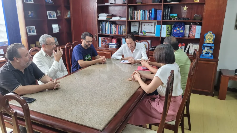 Fotografia colorida em plano médio de uma reunião institucional realizada em um gabinete oficial. Seis pessoas, cinco homens e uma mulher, estão sentadas em torno de uma mesa retangular de tampo de granito claro. O ambiente possui estantes de madeira ao fundo com livros e troféus, indicando um ambiente governamental ou administrativo. Os participantes estão em atitude de diálogo, com papéis e cadernos sobre a mesa, representando a articulação entre a ACIVA, a UFSC e a Prefeitura de Araranguá para a retomada das obras do Centro de Ciências da Saúde.