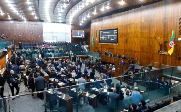 ALRS define hoje pauta com reajuste do magistério Foto em ângulo alto do Plenário 20 de Setembro da Assembleia Legislativa do Rio Grande do Sul (ALRS), em Porto Alegre, durante uma sessão plenária movimentada. Deputados estaduais estão sentados em suas bancadas de madeira, equipadas com laptops e monitores, enquanto outros circulam. A mesa diretora elevada está ao fundo sob uma grande tela de LED que exibe informações de votação e uma transmissão ao vivo do palestrante. A bandeira do RS e o brasão do estado são visíveis na parede de madeira. É o local onde ocorrem as votações decisivas de projetos de lei, como o reajuste do magistério estadual e a criação do Fundo de Bem-estar Animal, descritos na matéria.