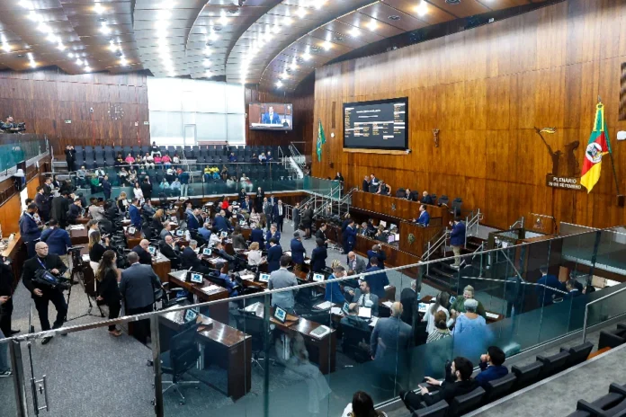 ALRS define hoje pauta com reajuste do magistério Foto em ângulo alto do Plenário 20 de Setembro da Assembleia Legislativa do Rio Grande do Sul (ALRS), em Porto Alegre, durante uma sessão plenária movimentada. Deputados estaduais estão sentados em suas bancadas de madeira, equipadas com laptops e monitores, enquanto outros circulam. A mesa diretora elevada está ao fundo sob uma grande tela de LED que exibe informações de votação e uma transmissão ao vivo do palestrante. A bandeira do RS e o brasão do estado são visíveis na parede de madeira. É o local onde ocorrem as votações decisivas de projetos de lei, como o reajuste do magistério estadual e a criação do Fundo de Bem-estar Animal, descritos na matéria.