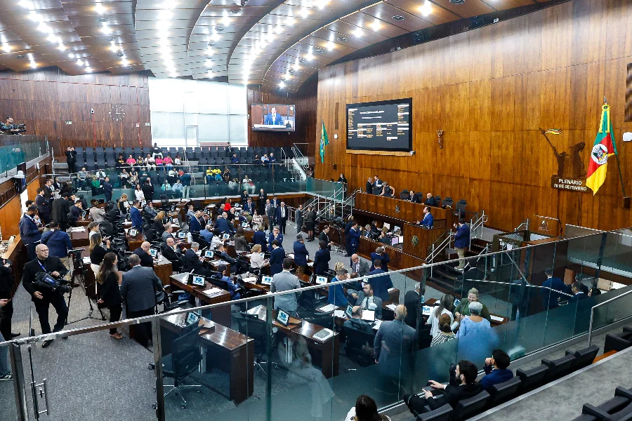 Foto em ângulo alto do Plenário 20 de Setembro da Assembleia Legislativa do Rio Grande do Sul (ALRS), em Porto Alegre, durante uma sessão plenária movimentada. Deputados estaduais estão sentados em suas bancadas de madeira, equipadas com laptops e monitores, enquanto outros circulam. A mesa diretora elevada está ao fundo sob uma grande tela de LED que exibe informações de votação e uma transmissão ao vivo do palestrante. A bandeira do RS e o brasão do estado são visíveis na parede de madeira. É o local onde ocorrem as votações decisivas de projetos de lei, como o reajuste do magistério estadual e a criação do Fundo de Bem-estar Animal, descritos na matéria.