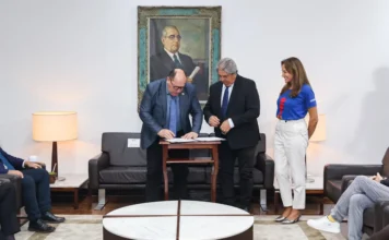 ALRS e Gramado renovam parceria para Prêmio de Cinema Foto jornalística no Gabinete da Presidência da ALRS, em Porto Alegre, capturando o momento exato da assinatura do acordo de cooperação para a 23ª Mostra de Curtas Gaúchos. No centro, o Presidente da ALRS, Deputado Sergio Peres, assina o documento na mesa de centro de madeira, observado atentamente pelo Prefeito de Gramado, Nestor Tissot (de terno escuro) e pela Presidente da Gramadotur, Rosa Helena Volker (de blusa azul e calça branca), que está em pé e sorrindo. O ambiente é formal, com um grande retrato histórico na parede, um sofá de couro e outras pessoas sentadas no primeiro plano. Esta imagem ilustra o compromisso institucional com o fomento do audiovisual regional no 54º Festival de Cinema de Gramado.