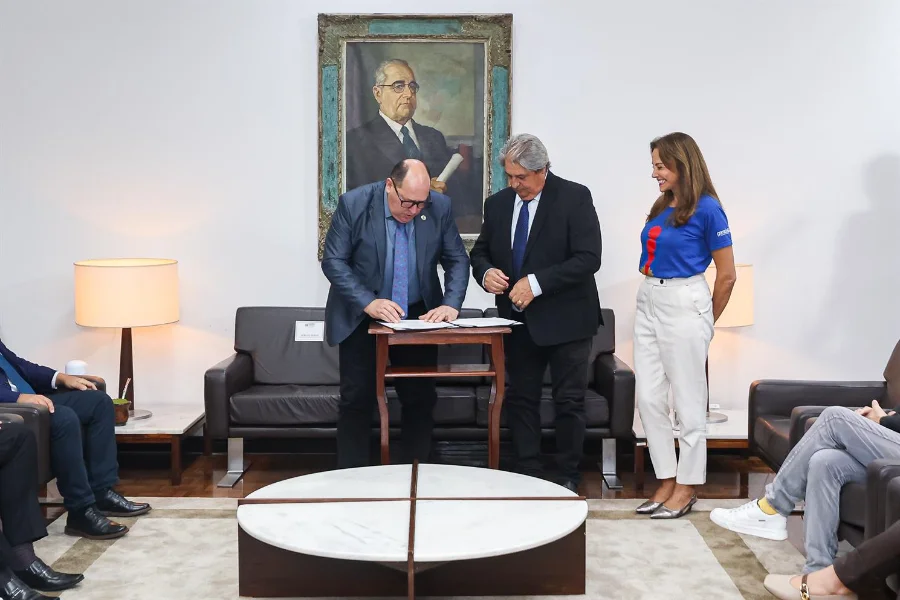 ALRS e Gramado renovam parceria para Prêmio de Cinema Foto jornalística no Gabinete da Presidência da ALRS, em Porto Alegre, capturando o momento exato da assinatura do acordo de cooperação para a 23ª Mostra de Curtas Gaúchos. No centro, o Presidente da ALRS, Deputado Sergio Peres, assina o documento na mesa de centro de madeira, observado atentamente pelo Prefeito de Gramado, Nestor Tissot (de terno escuro) e pela Presidente da Gramadotur, Rosa Helena Volker (de blusa azul e calça branca), que está em pé e sorrindo. O ambiente é formal, com um grande retrato histórico na parede, um sofá de couro e outras pessoas sentadas no primeiro plano. Esta imagem ilustra o compromisso institucional com o fomento do audiovisual regional no 54º Festival de Cinema de Gramado.